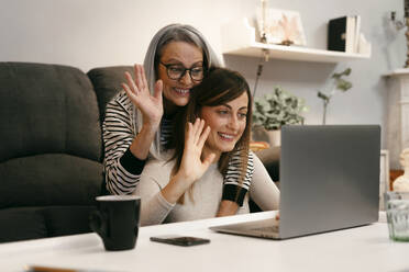 Happy women waving hand to video call on laptop while sitting at home - ERRF04761