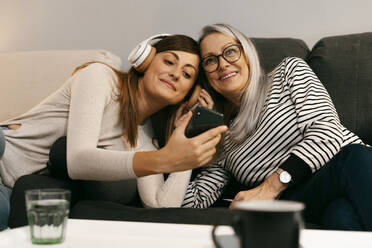 Woman with headphones showing mobile phone to senior woman while resting on sofa at home - ERRF04768