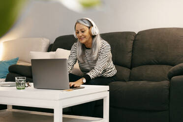 Smiling woman wearing headphones using laptop while sitting on sofa at home - ERRF04773