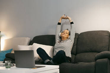 Carefree woman with hand raised listening music while sitting on sofa at home - ERRF04775