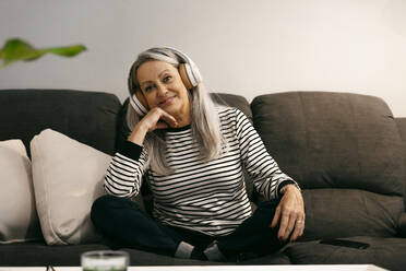 Senior woman wearing headphones smiling while sitting with hand on chin at home - ERRF04777