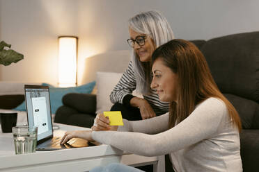 Lächelnde Mutter und Tochter beim Online-Shopping am Laptop, während sie zu Hause sitzen - ERRF04780