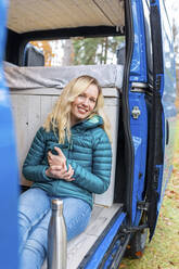 Lächelnde Frau mit Teetasse, die an der Tür eines Wohnmobils sitzt - WPEF03742