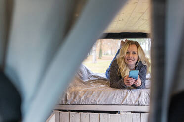 Lächelnde Frau auf dem Bett mit Handy in einem Lieferwagen - WPEF03750