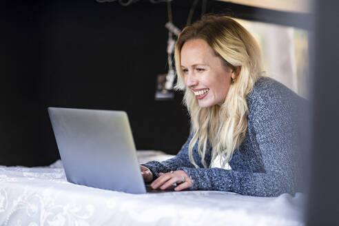 Glückliche Frau arbeitet am Laptop, während sie auf dem Bett im Wohnmobil liegt - WPEF03782