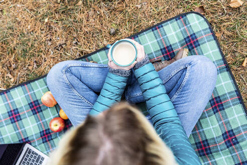 Woman holding tea cup while sitting on picnic blanket at remote location - WPEF03790