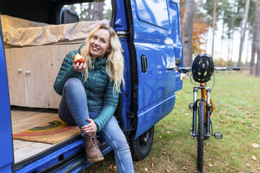 Lächelnde Frau, die einen Apfel isst, während sie an der Tür eines Wohnmobils in Cannock Chase sitzt - WPEF03799