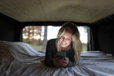 Lächelnde Frau mit blondem Haar liegt auf dem Bett und benutzt ihr Smartphone im Wohnmobil - WPEF03805