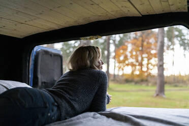 Comfortable woman looking outside while lying on bed in camper van - WPEF03814