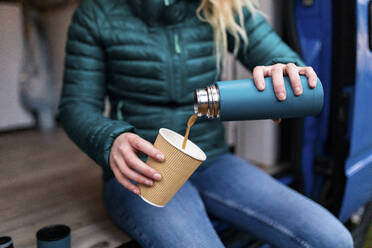 Mid adult woman pouring tea in disposable cup at door of camper van - WPEF03822