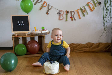 Lächelnder kleiner Junge sitzt mit Kuchen auf dem Boden gegen Dekoration zu Hause - NGF00723