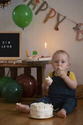 Hungriger kleiner Junge, der den Kuchen seines ersten Geburtstags isst, während er gegen die Geburtstagsdekoration zu Hause sitzt - NGF00726