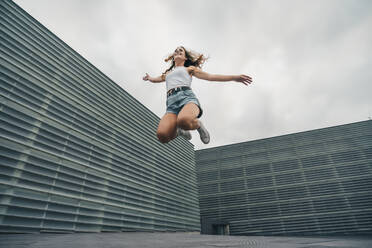 Carefree young woman jumping with arms outstretched against sky - MTBF00731