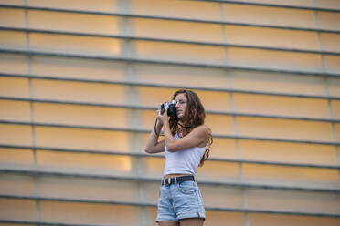 Woman taking photo through camera while standing against wall - MTBF00741