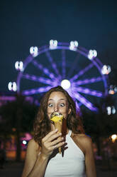 Woman looking at ice cream while standing against ferries wheel in city - MTBF00744