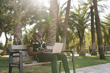 Männlicher Unternehmer hört Musik, während er im Park am Laptop arbeitet - EGAF01209