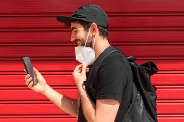 Side view of unshaven male in face mask on cellphone during video chat with girlfriend on street near red wall - ADSF18608