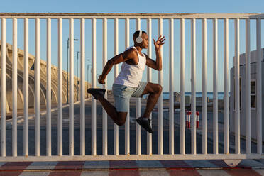 Full body side view of concentrated African American runner listening to music in headphones while jumping above ground during jogging training - ADSF18614