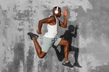 Full body side view of concentrated African American runner listening to music in headphones while jumping above ground during jogging training - ADSF18615