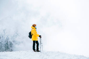 Seitenansicht eines aktiven Reisenden in leuchtend gelber warmer Jacke mit Rucksack, der Trekkingstöcke und eine medizinische Maske hält, während er auf einem verschneiten Hügel steht und die Landschaft bewundert - ADSF18621