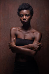 Sinnliche afroamerikanische Frau in engem schwarzen Top mit nackten Schultern, die mit verschränkten Armen in einem dunklen Studio steht und provokativ in die Kamera schaut - ADSF18646