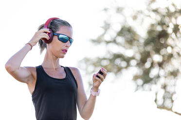 Konzentrierte junge Frau in schwarzem Hemd und Sonnenbrille, die einen modernen MP3-Player benutzt und über Kopfhörer Musik hört, während sie einen sonnigen Tag im Park verbringt - ADSF18675