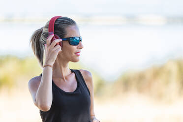 Schlanke Sportlerin in Sportkleidung und Sonnenbrille, die sich vor dem Sport im sonnigen Park ein Headset aufsetzt - ADSF18677