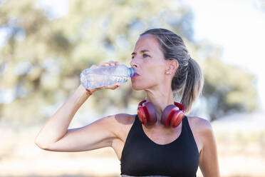 Ruhige Sportlerin im Sport-BH und mit Kopfhörern, die frisches Wasser aus einer Flasche trinkt, während sie sich nach dem Training an einem sonnigen Tag entspannt und wegschaut - ADSF18679