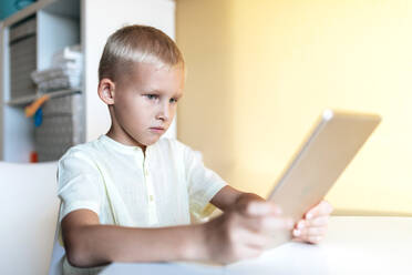 Seitenansicht des konzentrierten blondhaarigen kleinen Jungen im weißen Hemd beim Surfen im Internet auf einem Tablet in einem hellen Raum im Sonnenlicht - ADSF18680