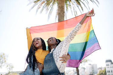 Niedriger Winkel von entzückten, verliebten afroamerikanischen Frauen, die sich auf der Straße umarmen, während sie mit einer LGBT-Regenbogenfahne stehen und wegschauen - ADSF18692