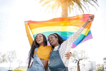 Niedriger Winkel von entzückten, verliebten afroamerikanischen Frauen, die sich auf der Straße umarmen, während sie mit einer LGBT-Regenbogenfahne stehen und wegschauen - ADSF18693