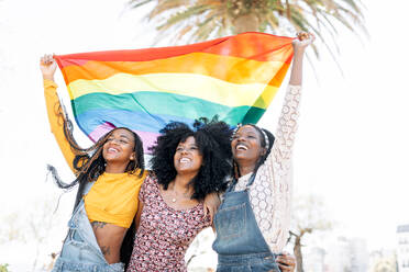 Niedriger Winkel von entzückten, verliebten afroamerikanischen Frauen, die sich auf der Straße umarmen, während sie mit einer LGBT-Regenbogenfahne stehen und wegschauen - ADSF18695