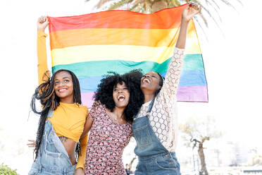 Niedriger Winkel von entzückten, verliebten afroamerikanischen Frauen, die sich auf der Straße umarmen, während sie mit einer LGBT-Regenbogenfahne stehen und wegschauen - ADSF18696