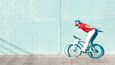 Side view of young active bearded male cyclist in stylish outfit listening to music in headphones while riding bike on street near blue stone building on sunny day - ADSF18707