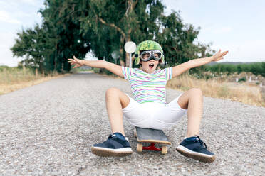 Ebenerdiges, zufriedenes Kind mit Wassermelonenhelm und Schutzbrille, das auf einem Skateboard auf einer asphaltierten Fahrbahn sitzt und wegschaut - ADSF18709