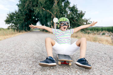 Ground level of content kid in watermelon helmet and goggles sitting on skateboard on asphalt roadway and looking away - ADSF18709