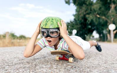 Ebenerdiges Bild eines glücklichen Kindes mit Schutzbrille und dekorativem Wassermelonenhelm, das auf einem Skateboard auf der Fahrbahn liegt und wegschaut - ADSF18711