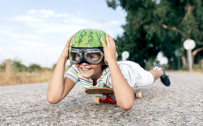 Ebenerdiges Bild eines glücklichen Kindes mit Schutzbrille und dekorativem Wassermelonenhelm, das auf einem Skateboard auf der Fahrbahn liegt und wegschaut - ADSF18712