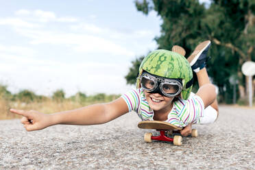 Ground level of happy kid in protective eyewear and ornamental watermelon helmet lying on skateboard on roadway and looking away - ADSF18716