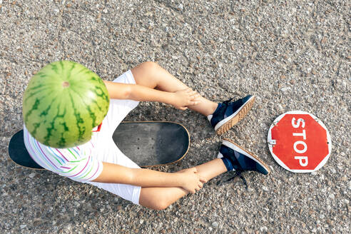Stylish Kind in ornamentalen Wassermelone Kopf tragen und Sonnenbrille sitzt auf Skateboard in der Nähe Schild mit STOP Titel auf der Straße - ADSF18717