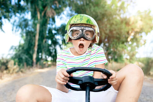 Niedriger Winkel des erstauntes Kind in modischer Kleidung und Schutzbrille fahren Go-Kart mit Pedalen auf der Fahrbahn, während wegschauen - ADSF18718
