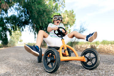 Low angle of amazed kid in trendy wear and goggles riding go kart with pedals on roadway while looking away - ADSF18720