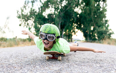 Ebenerdiges Bild eines glücklichen Kindes mit Schutzbrille und dekorativem Wassermelonenhelm, das auf einem Skateboard auf der Fahrbahn liegt und wegschaut - ADSF18725