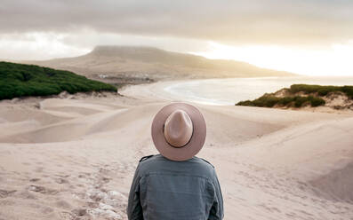 Back view unrecognizable male in denim jacket and hat standing on sandy beach and admiring picturesque hilly seacoast view - ADSF18733