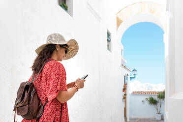 Back view of cheerful female traveler in straw hat and sunglasses text messaging on cellphone near old house in White Village - ADSF18746