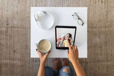 Von oben auf eine nicht erkennbare Frau, die sich ein Video auf einem Tablet ansieht, während sie mit einer Tasse Kaffee am Tisch sitzt und das Wochenende zu Hause verbringt - ADSF18758