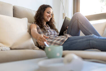 Positive female wearing domestic clothes sitting on soft couch at home and chatting on social media with friends via tablet - ADSF18760