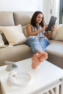 Positive Frau in häuslicher Kleidung, die zu Hause auf einer weichen Couch sitzt und mit Freunden über ein Tablet in sozialen Medien chattet - ADSF18763