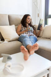 Positive female wearing domestic clothes sitting on soft couch at home and chatting on social media with friends via tablet - ADSF18764