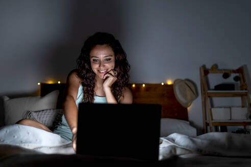 Delighted female in nightwear lying on bed in dark room and watching funny video on netbook while smiling and spending weekend at home - ADSF18765